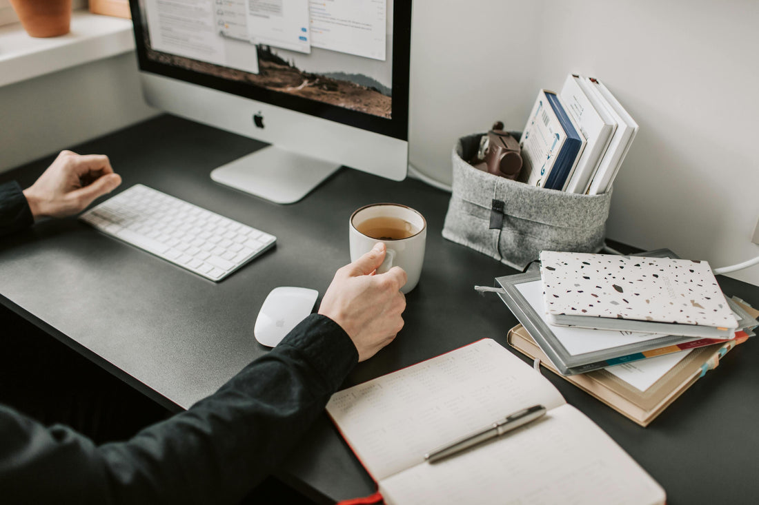 hands of a focused man holding a cup of coffee on a work desk with a desktop computer and notebooks