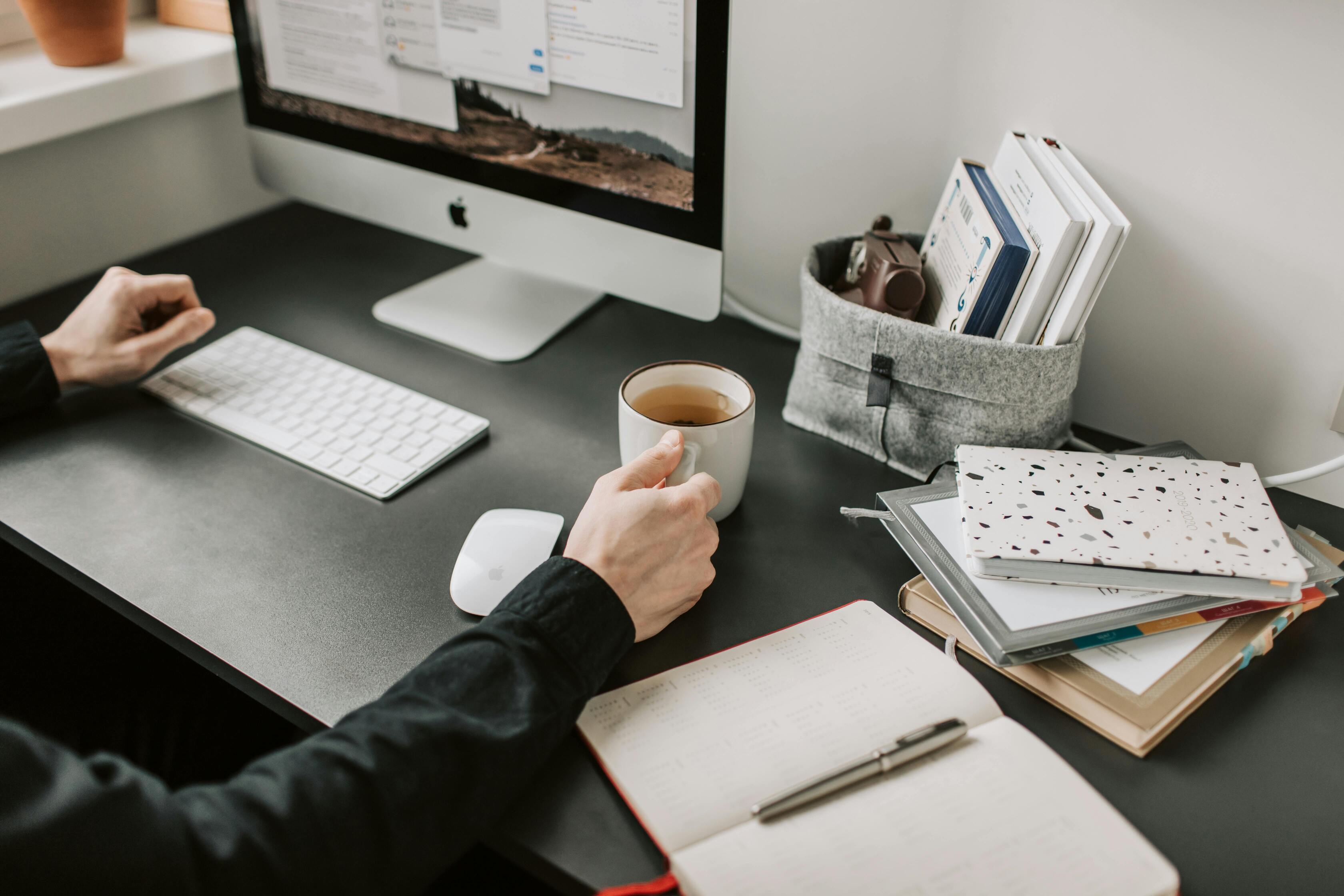 hands of a focused man holding a cup of coffee on a work desk with a desktop computer and notebooks