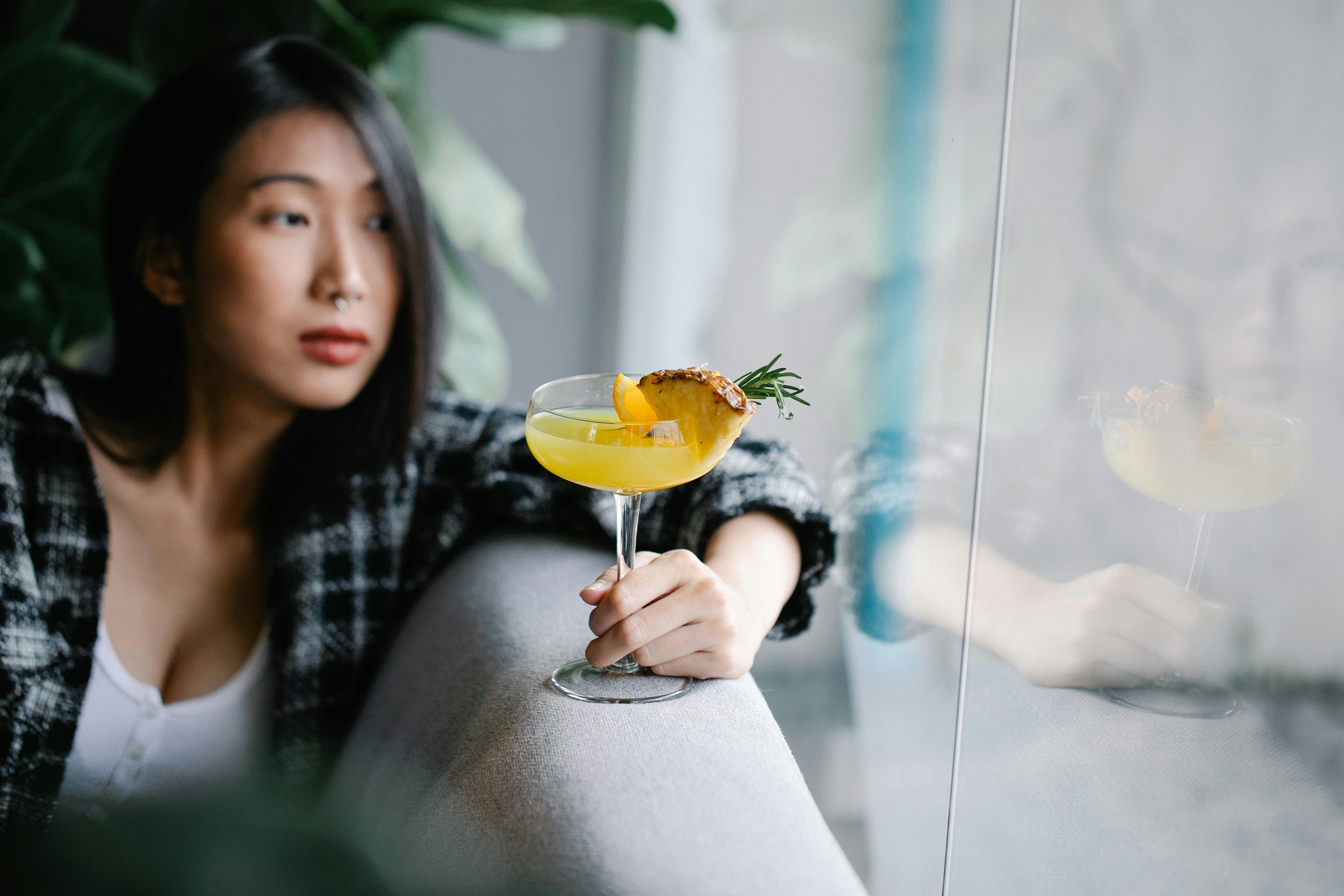 Asian woman leaning on a couch looking outside a window holding a pineapple mocktail